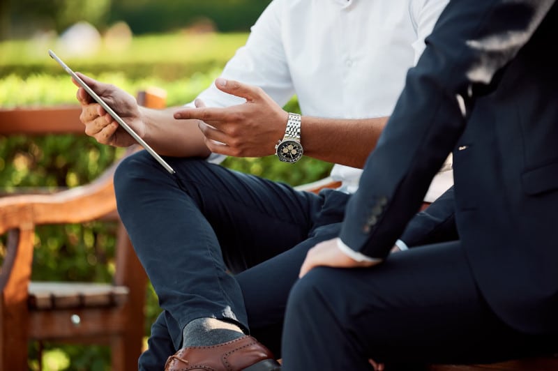 two well-dressed men sitting outside; one is consulting the other one and showing them something on a tablet computer or smartphone