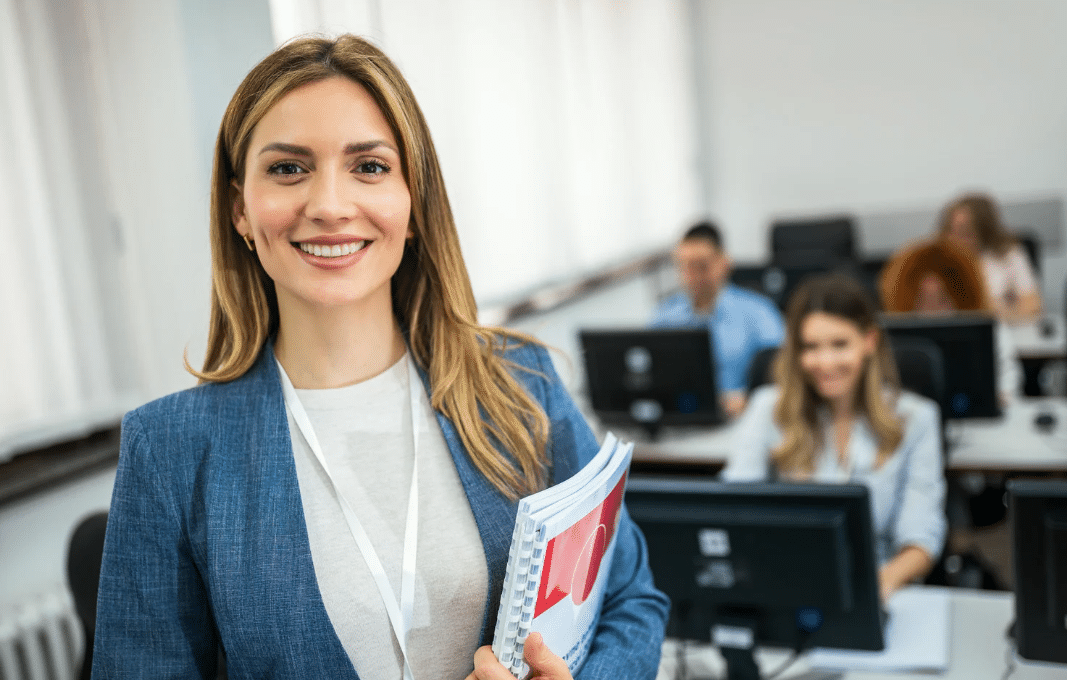 woman smiling while holding stack of papers with people on computers in the background