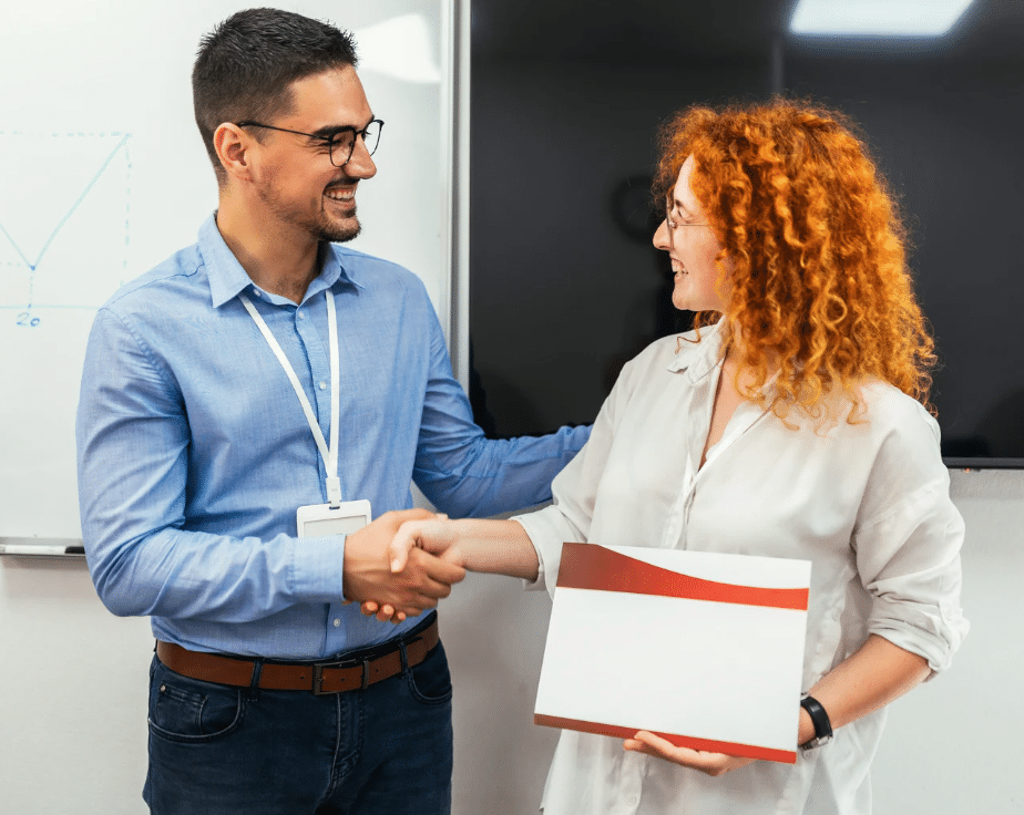 man shaking hands with woman who is holding a certificate, in front of a blackboard in a classroom